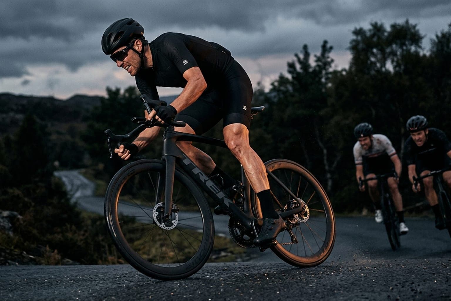 Road cyclist launching an explosive out-of-the-saddle attack over a dark asphalt crest ahead of two chasing riders.