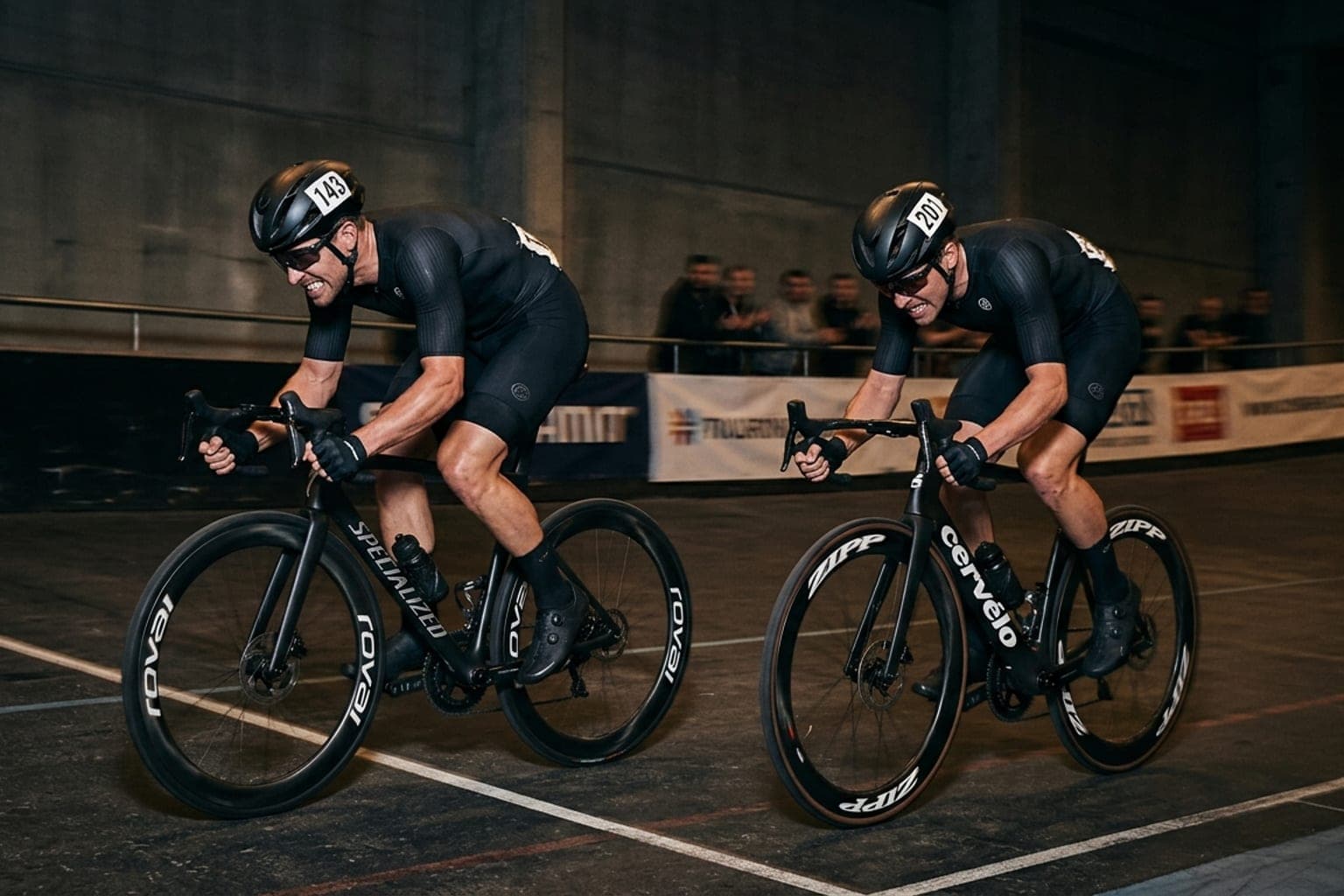 Two road cyclists tucked low in the drops sprinting head-to-head toward a finish line in a dark concrete tunnel.